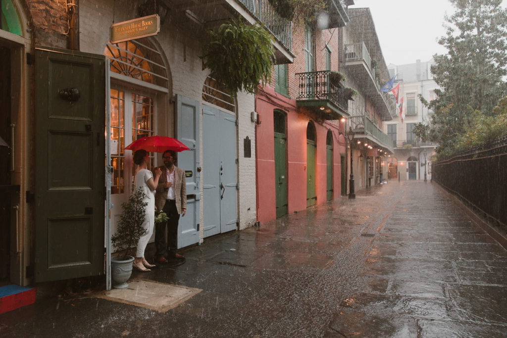 Couple stands in Pirate's Alley under a red umbrella during their stormy French Quarter elopement.